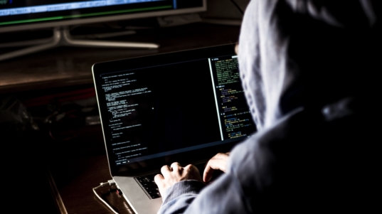 hacker in hoodie typing code in program using laptop in dark studio